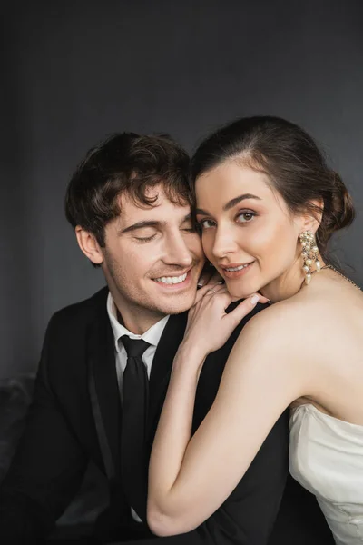 Portrait of happy and young brunette groom in black suit smiling near pretty bride in luxurious earrings with pearls and white wedding dress in hotel room — Stock Photo