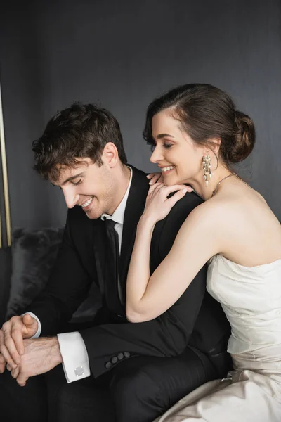 Retrato de encantadora novia joven en vestido de novia blanco y novio guapo en traje negro sonriendo juntos mientras están sentados en la habitación de hotel, felices recién casados - foto de stock