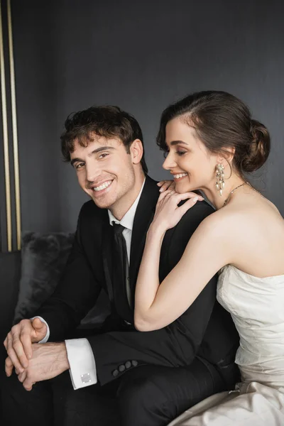 Портрет очаровательной невесты в белом свадебном платье, улыбающейся рядом с красивым женихом в черном костюме, смотрящим в камеру, сидя в гостиничном номере, счастливые молодожены — стоковое фото