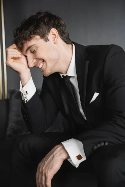 Portrait of joyful and young groom in black suit with white shirt and tie touching face with hand while smiling and sitting on comfortable couch in modern hotel room on wedding day — Stock Photo