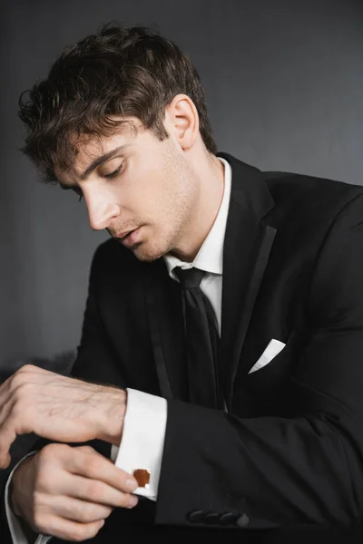 Retrato de novio guapo y joven en traje negro con camisa blanca y corbata ajustando gemelos y sentado en cómodo sofá gris oscuro en la habitación de hotel moderna el día de la boda - foto de stock