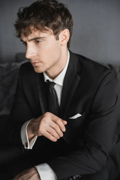Retrato de boa aparência jovem noivo em terno preto com camisa branca e gravata sentado no confortável sofá cinza escuro no quarto de hotel moderno no dia do casamento — Fotografia de Stock