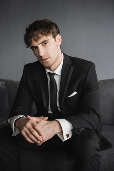 Portrait of good looking young groom in black suit with white shirt and tie sitting on comfortable dark grey couch and looking at camera in modern hotel room on wedding day — Stock Photo