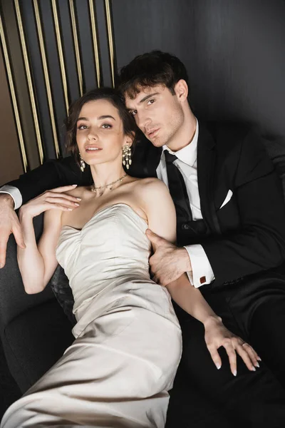 Good looking groom in black suit with tie hugging charming young bride in luxurious earrings with pearls and necklace posing in white wedding dress on dark grey couch in hotel room — Stock Photo