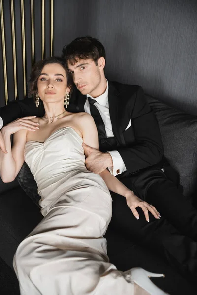 Charming young bride in luxurious earrings with pearls and white wedding dress and good looking groom in black suit with tie sitting together on dark grey couch in hotel room and looking at camera — Stock Photo