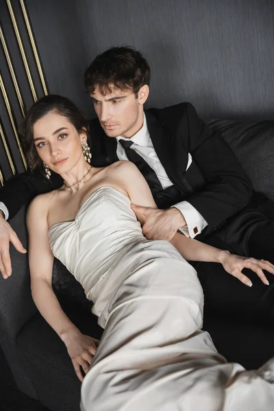 Charming young bride in luxurious earrings with pearls and white wedding dress leaning on groom in black suit with tie while sitting together on dark grey couch in hotel room — Stock Photo