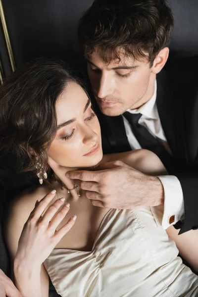 Overhead view of groom in black suit with tie touching face of charming young bride in white wedding dress and luxurious jewelry while sitting together in hotel room — Stock Photo