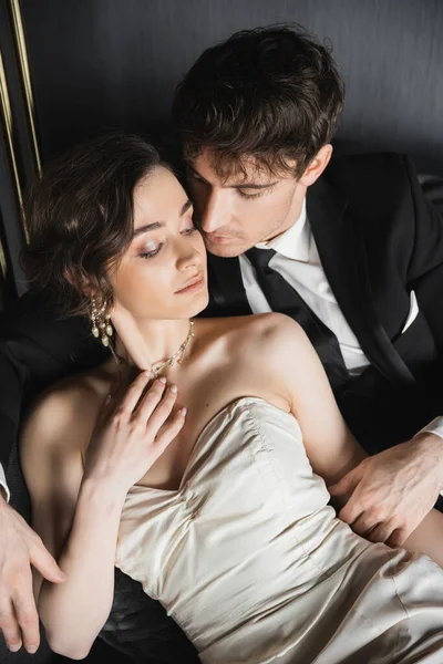 Overhead view of groom in black suit with tie hugging charming bride in earrings with pearls touching her necklace and sitting in white wedding dress on dark grey couch in hotel room — Stock Photo