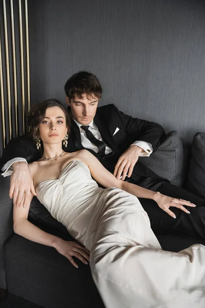 Charming young bride in earrings with pearls and white wedding dress posing next to good looking groom in black suit with tie while lying together on dark grey couch in hotel room — Stock Photo