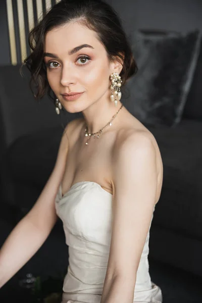 Porträt einer charmanten jungen Braut mit brünetten Haaren im eleganten weißen Hochzeitskleid, luxuriösem Schmuck, Ohrringen mit Perlen und Halskette, die im Hotelzimmer in die Kamera schauen — Stockfoto