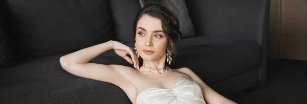 Portrait of dreamy and gorgeous bride with brunette hair sitting in elegant and white wedding dress, luxurious jewelry, earrings and necklace and looking away in hotel room, banner — Stock Photo
