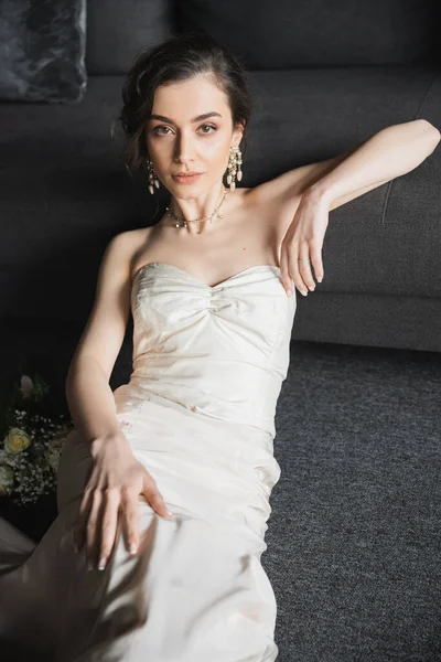 Encantador jovem noiva com cabelo morena sentado em vestido de noiva elegante e branco, jóias de luxo, brincos e colar olhando para a câmera perto de buquê de noiva no quarto de hotel — Fotografia de Stock