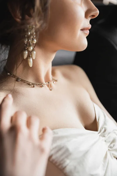 Close up view of bride with brunette hair posing in elegant and white wedding dress, luxurious jewelry, earrings with pearls and necklace in hotel room, details of wedding accessories — Stock Photo