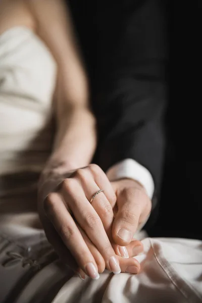 Vista recortada de los recién casados felices, novia con elegante y lujoso anillo de bodas en el dedo y el novio tomados de la mano uno del otro después de la boda en la habitación de hotel - foto de stock