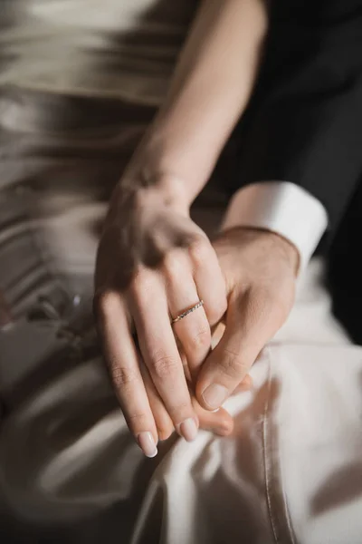 Vista recortada de los recién casados, novia con elegante y lujoso anillo de bodas en el dedo y el novio en traje tomados de la mano el uno del otro después de la boda en la habitación de hotel - foto de stock