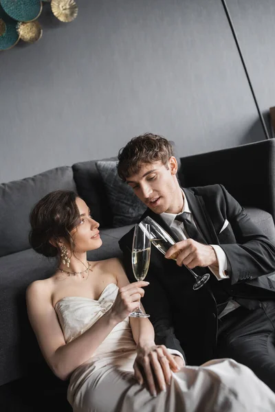Happy newlyweds, bride in white wedding dress and groom in black suit holding glasses of champagne while clinking and celebrating their marriage after wedding in hotel room — Stock Photo