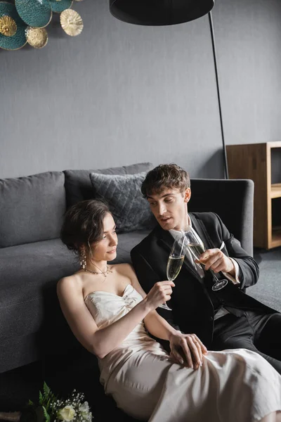 Bride in elegant white wedding dress and groom in black suit clinking glasses of champagne while celebrating their marriage near bridal bouquet after wedding in hotel room — Stock Photo