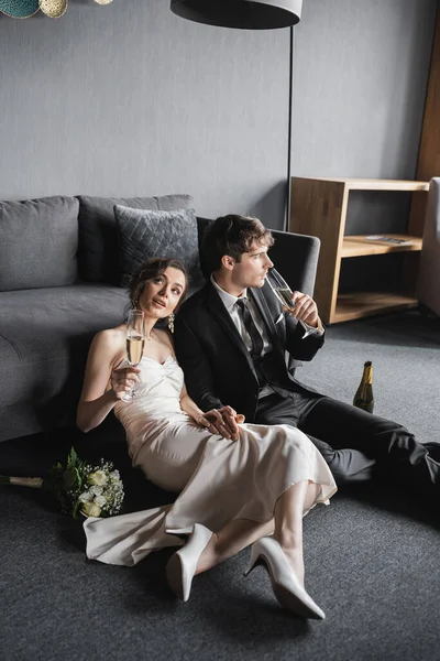 Dreamy bride in elegant white wedding dress and groom in black suit holding glasses of champagne while celebrating their marriage near bridal bouquet and couch after wedding in hotel room — Stock Photo