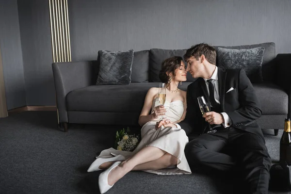 Feliz recém-casados, noiva em vestido de noiva branco e noivo em terno preto segurando copos de champanhe ao beijar e celebrar seu casamento perto de buquê de noiva e sofá no quarto de hotel — Fotografia de Stock