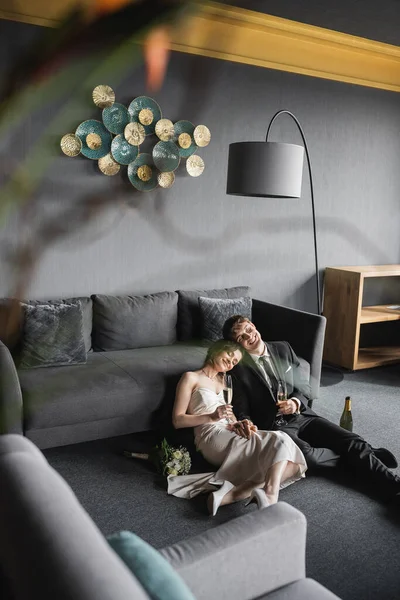 Glückliche Brautpaare mit geschlossenen Augen, die Champagnergläser halten, während sie ihre Hochzeit in der Nähe von Brautstrauß und Flasche auf dem Boden feiern, nach der Hochzeit im Hotelzimmer mit Couch und Stehlampe — Stockfoto