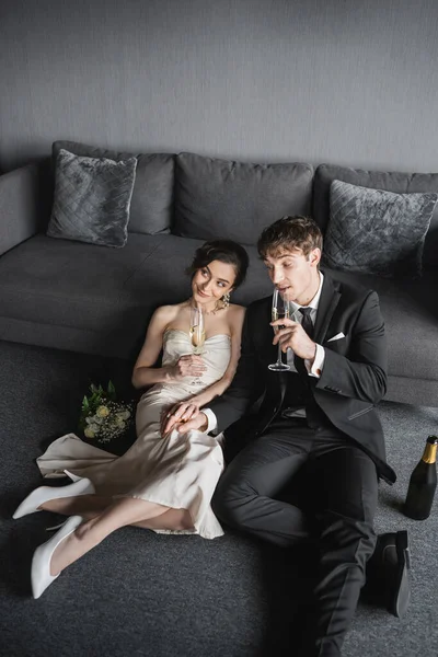 Happy bride in white wedding dress and groom in black suit holding glasses of champagne while celebrating their marriage near bridal bouquet and bottle after wedding in hotel room with couch — Stock Photo