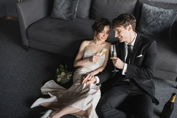 Счастливые молодожены в элегантной одежде звон бокалов шампанского во время празднования их брака возле свадебного букета и бутылки на полу после свадьбы в номере отеля с диваном — стоковое фото