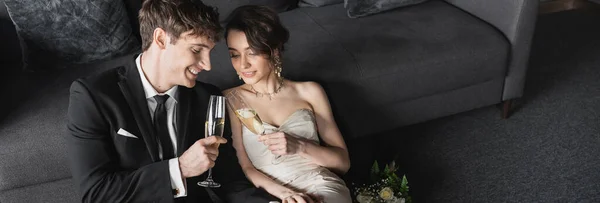 Happy bride in white wedding dress and groom in black suit clinking glasses of champagne while celebrating their marriage near bridal bouquet after wedding in hotel room, banner — Stock Photo