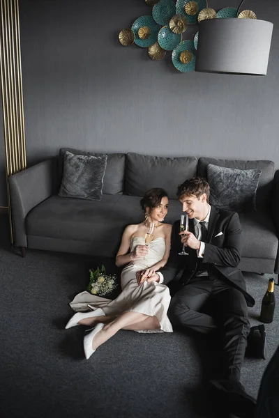 Cheerful bride in white dress and groom in black suit holding glasses of champagne while celebrating their marriage near bridal bouquet and couch after wedding in hotel room — Stock Photo