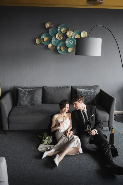 Fröhliche Braut im Brautkleid und Bräutigam im schwarzen Anzug trinken Champagner, während sie ihre Hochzeit in der Nähe von Brautstrauß, Couch und Stehlampe im Hotelzimmer feiern — Stockfoto