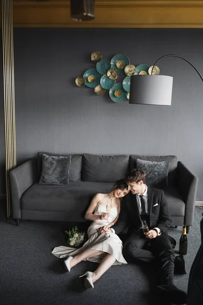 Noiva alegre em vestido de noiva apoiando-se no ombro do noivo em terno preto e segurando copos de champanhe perto de buquê de noiva, sofá e lâmpada de assoalho no quarto do hotel — Fotografia de Stock