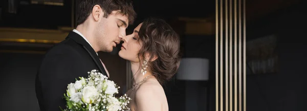 Side view of good looking groom in black formal wear kissing brunette bride with luxurious jewelry holding bridal bouquet of flowers while standing in hotel lobby, banner — Stock Photo