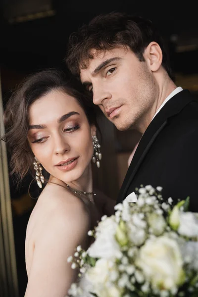 Good looking groom in black formal wear standing with brunette and elegant bride with makeup and jewelry holding bridal bouquet while standing in hotel lobby — Stock Photo
