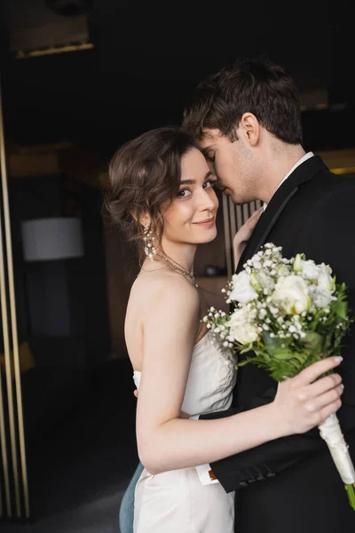 Noiva morena em vestido de noiva elegante segurando buquê de noiva com flores e abraçando o noivo em desgaste formal preto enquanto está de pé no lobby do hotel — Fotografia de Stock