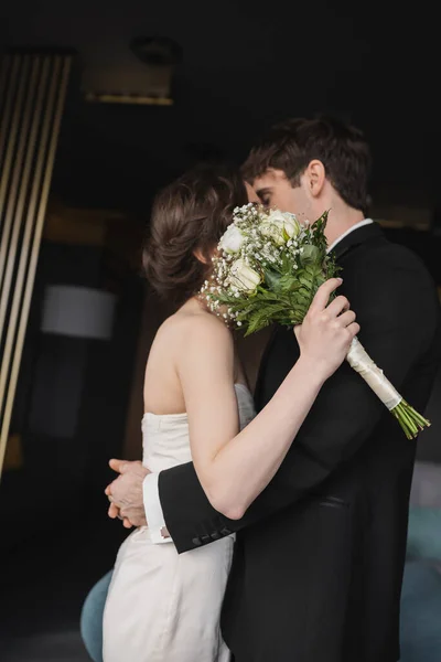 Брюнетка наречена в елегантній весільній сукні тримає весільний букет з квітами і обличчями, цілуючись з нареченим в чорному формальному одязі, стоячи в готельному лобі — Stock Photo
