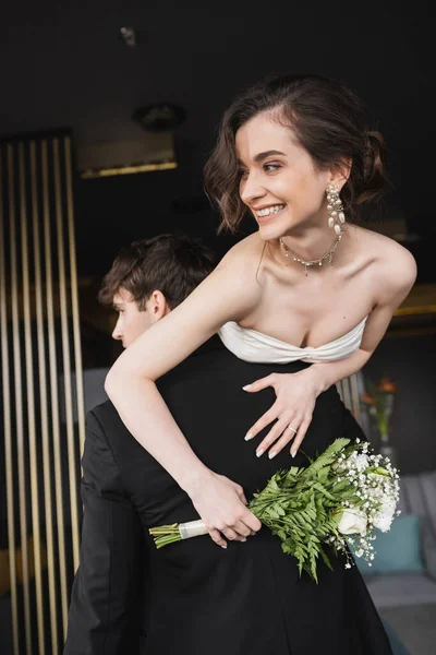 Groom in black formal wear lifting joyful bride in white wedding dress and luxurious jewelry holding bridal bouquet with flowers while standing in hotel lobby — Stock Photo