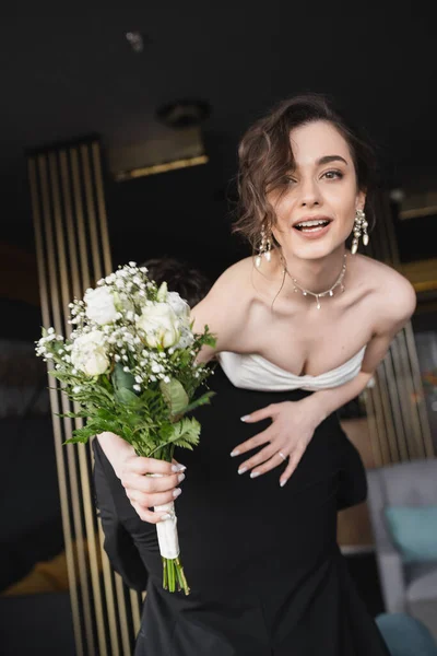 Noivo em preto formal desgaste levantando noiva bonita em vestido de noiva branco e jóias de luxo segurando buquê de noiva com flores, enquanto em pé no lobby do hotel — Fotografia de Stock