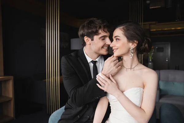Happy groom in black suit with tie touching face of charming bride in white wedding dress and jewelry while looking at each other in modern hotel suite — Stock Photo
