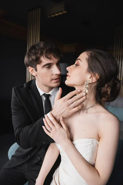 Groom in black suit with tie touching face of charming bride in white wedding dress and jewelry while looking at each other in hotel room, couple on wedding day — Stock Photo