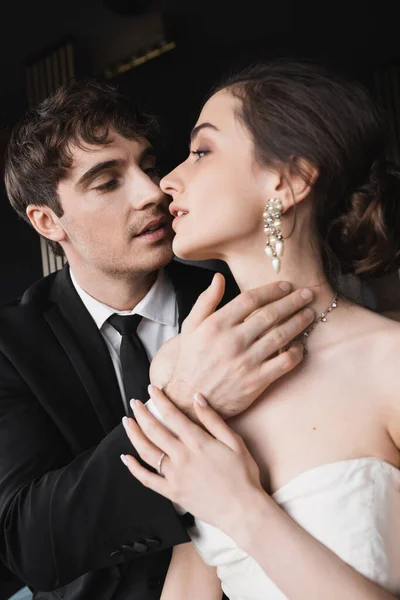 Portrait of tender groom in black suit with tie touching face of charming bride in white wedding dress and jewelry while looking at each other in hotel room — Stock Photo