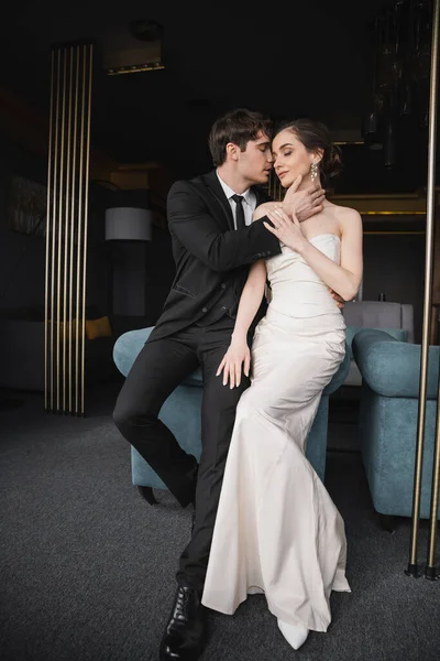 Zarter Bräutigam im schwarzen Anzug mit Krawatte berührt Gesicht der charmanten Braut im weißen Hochzeitskleid und Schmuck lehnt auf blauem Sofa im modernen Hotelzimmer — Stockfoto