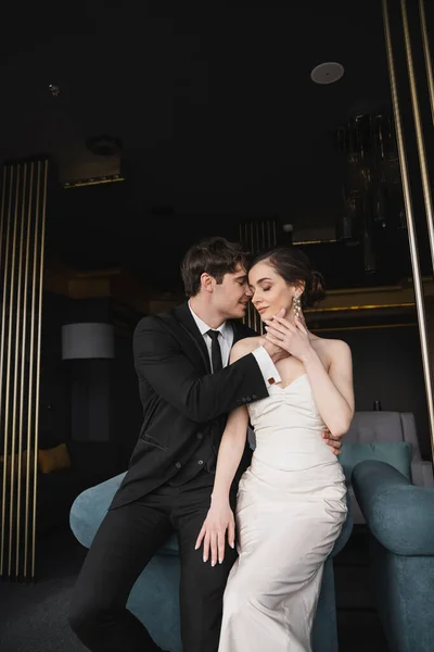 Happy groom in black suit with tie touching face of charming bride in white wedding dress and jewelry leaning on blue couch in modern hotel room — Stock Photo
