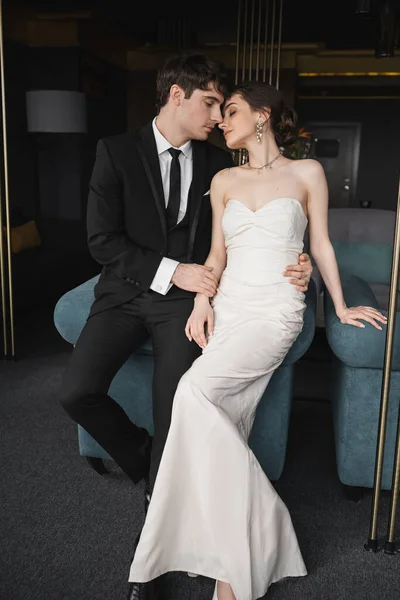 Zarter Bräutigam im schwarzen Anzug umarmt Taille der charmanten Braut in weißem Hochzeitskleid und Schmuck, während sie sich zusammen auf blauem Sofa im modernen Hotelzimmer lehnt — Stockfoto
