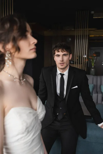 Handsome groom in black suit looking at camera while standing near charming bride in white dress and jewelry on blurred foreground in hotel room, couple on wedding day — Stock Photo