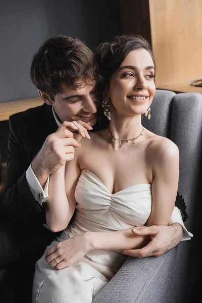Щасливий наречений в чорному костюмі тримає руку веселої нареченої в білій сукні і розкішні прикраси, посміхаючись і сидячи разом на зручному кріслі в готельному номері — стокове фото