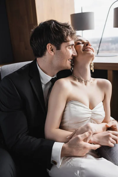 Happy groom in black suit hugging cheerful bride in white dress and luxurious jewelry while smiling and sitting together on comfortable armchair in hotel room — Stock Photo
