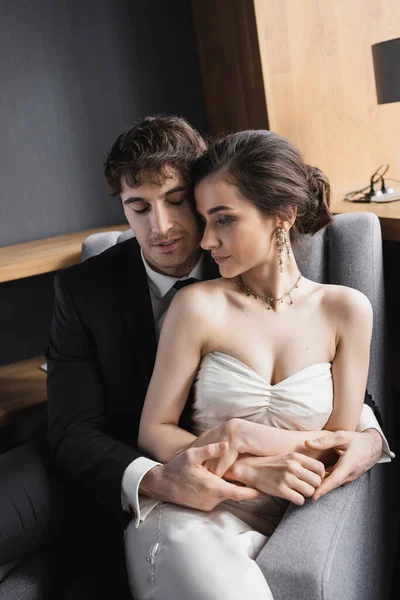 Наречений в чорному костюмі тримає руки ніжної нареченої в білій сукні і розкішні прикраси, сидячи разом на зручному кріслі в готельному номері, пара на день весілля — стокове фото