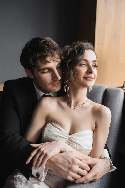 Good looking groom in black suit embracing charming bride in white dress and luxurious jewelry while smiling and sitting together on comfortable armchair in hotel room — Stock Photo