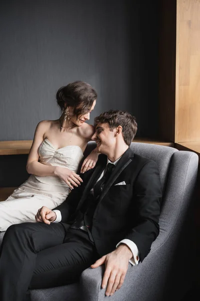 Брюнетка невеста в белом свадебном платье и роскошные украшения сидя вместе с счастливым женихом в черном костюме с галстуком на удобном кресле в гостиничном номере — стоковое фото