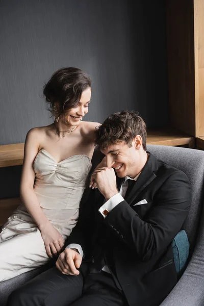 Noiva feliz em vestido de noiva branco e jóias luxuosas sorrindo e sentado junto com o noivo positivo em terno preto em poltrona confortável no quarto do hotel — Fotografia de Stock