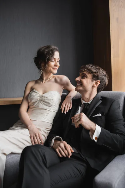 Glückliche Braut im weißen Hochzeitskleid und luxuriöser Halskette lächelnd, während sie zusammen mit positivem Bräutigam im schwarzen Anzug auf einem bequemen Sessel im Hotelzimmer sitzt — Stockfoto
